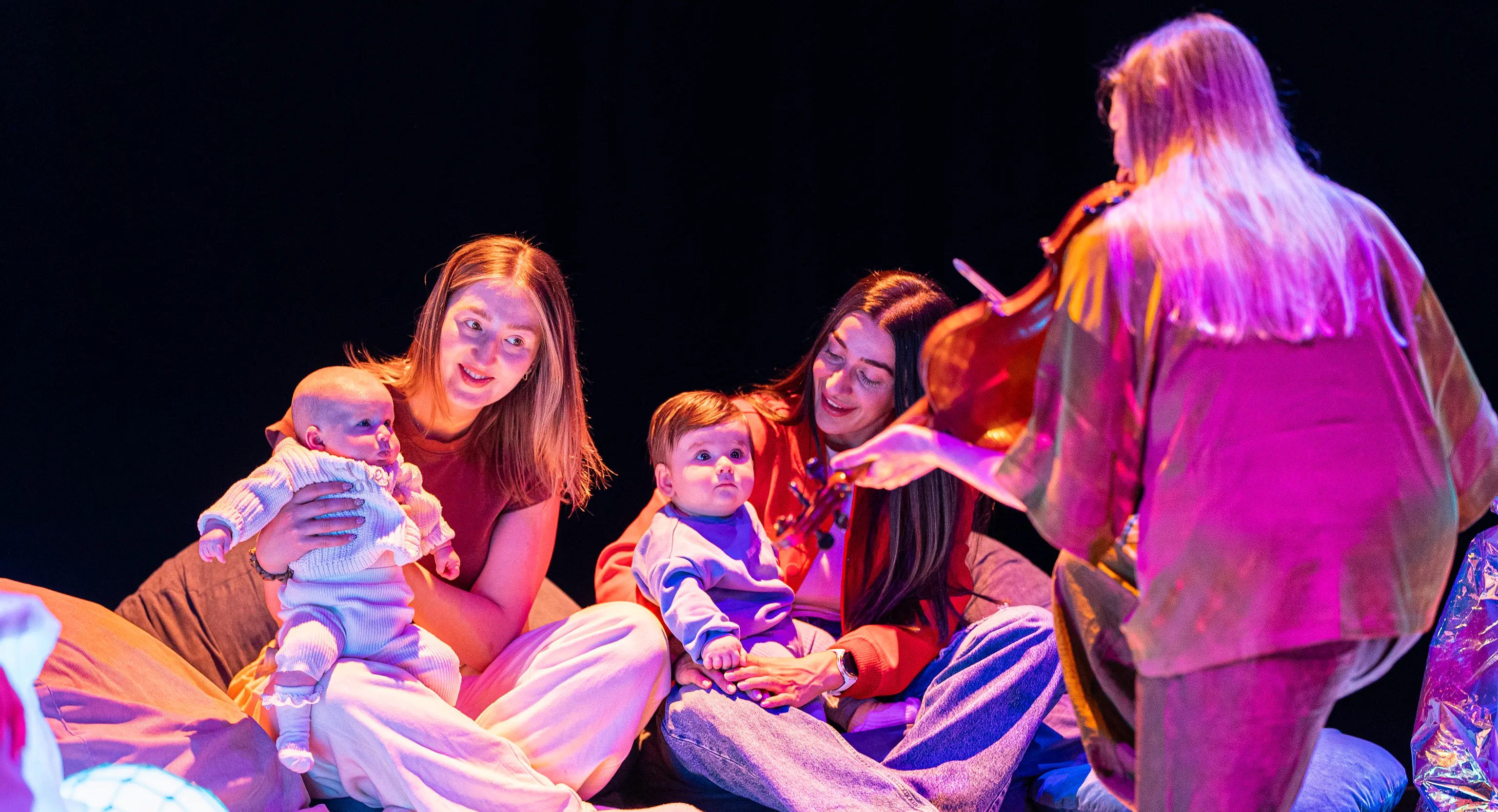 a group of parents/carers and their babies engage with a performer playing violin.