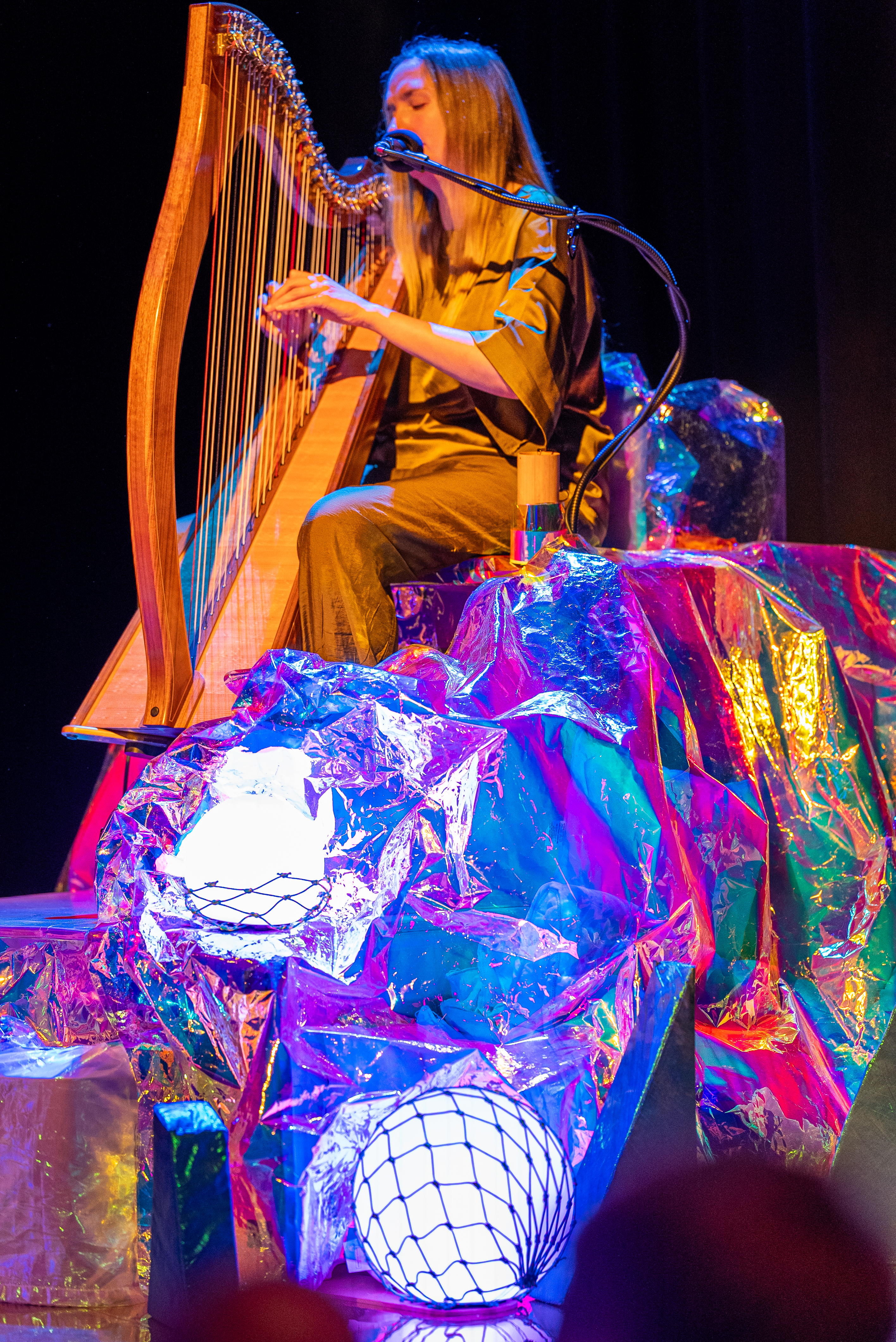 In the foreground are some props, fabric, spherical lights and netting. In background a person sits playing the harp.