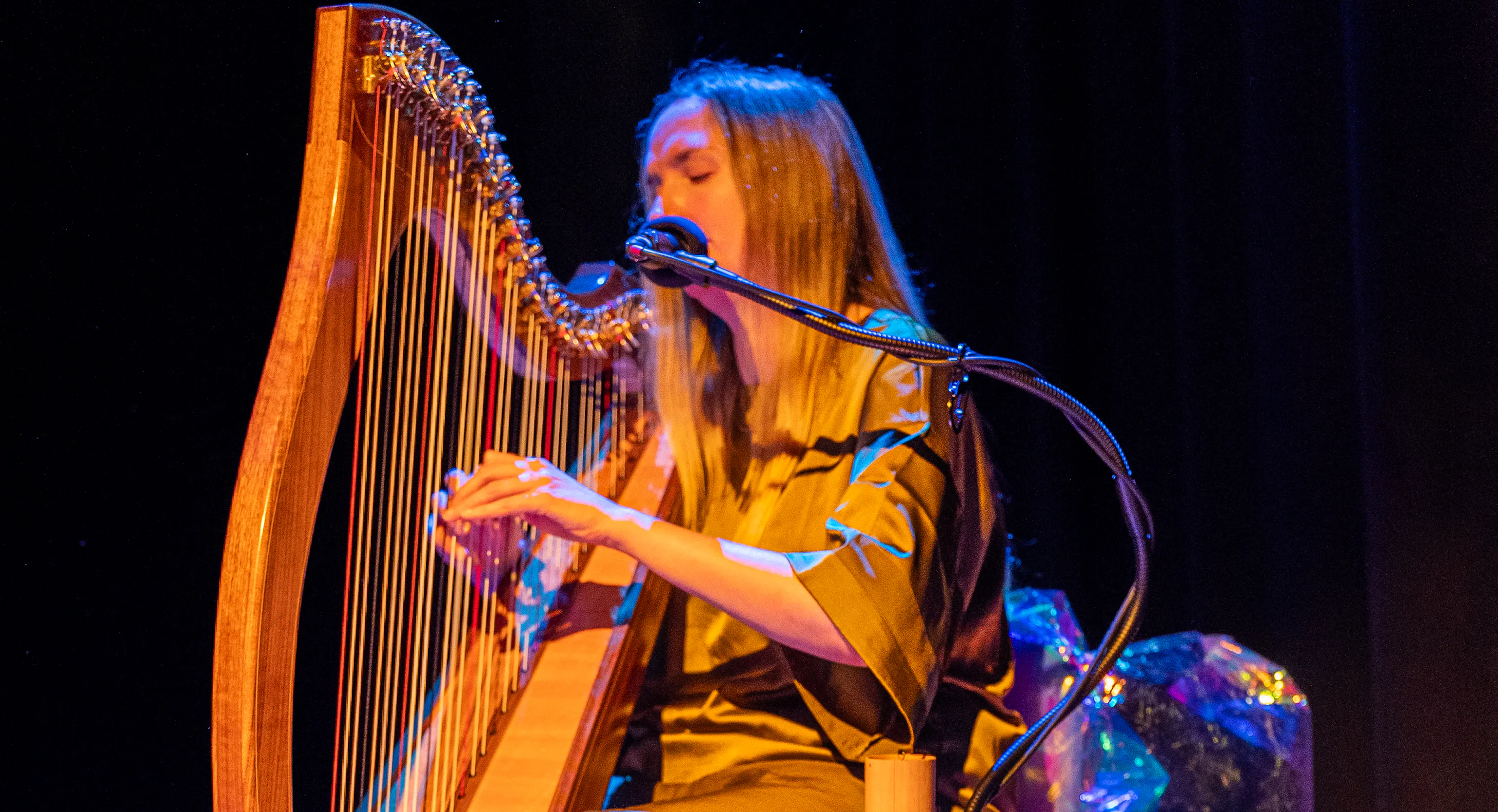 In the foreground are some props, fabric, spherical lights and netting. In background a person sits playing the harp.