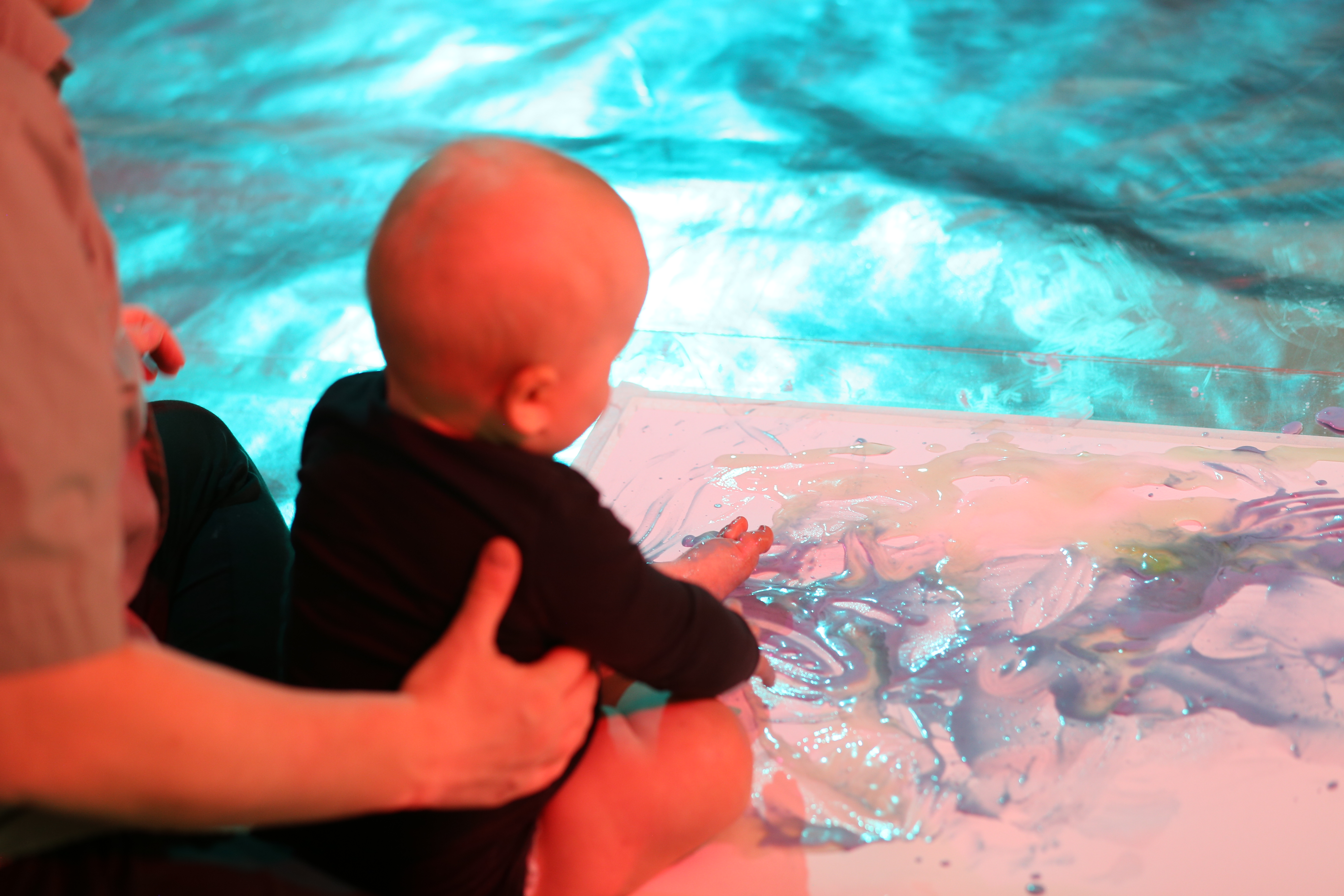 A parent holds a baby whilst they rub their feet in a gel like material on a piece of white paper