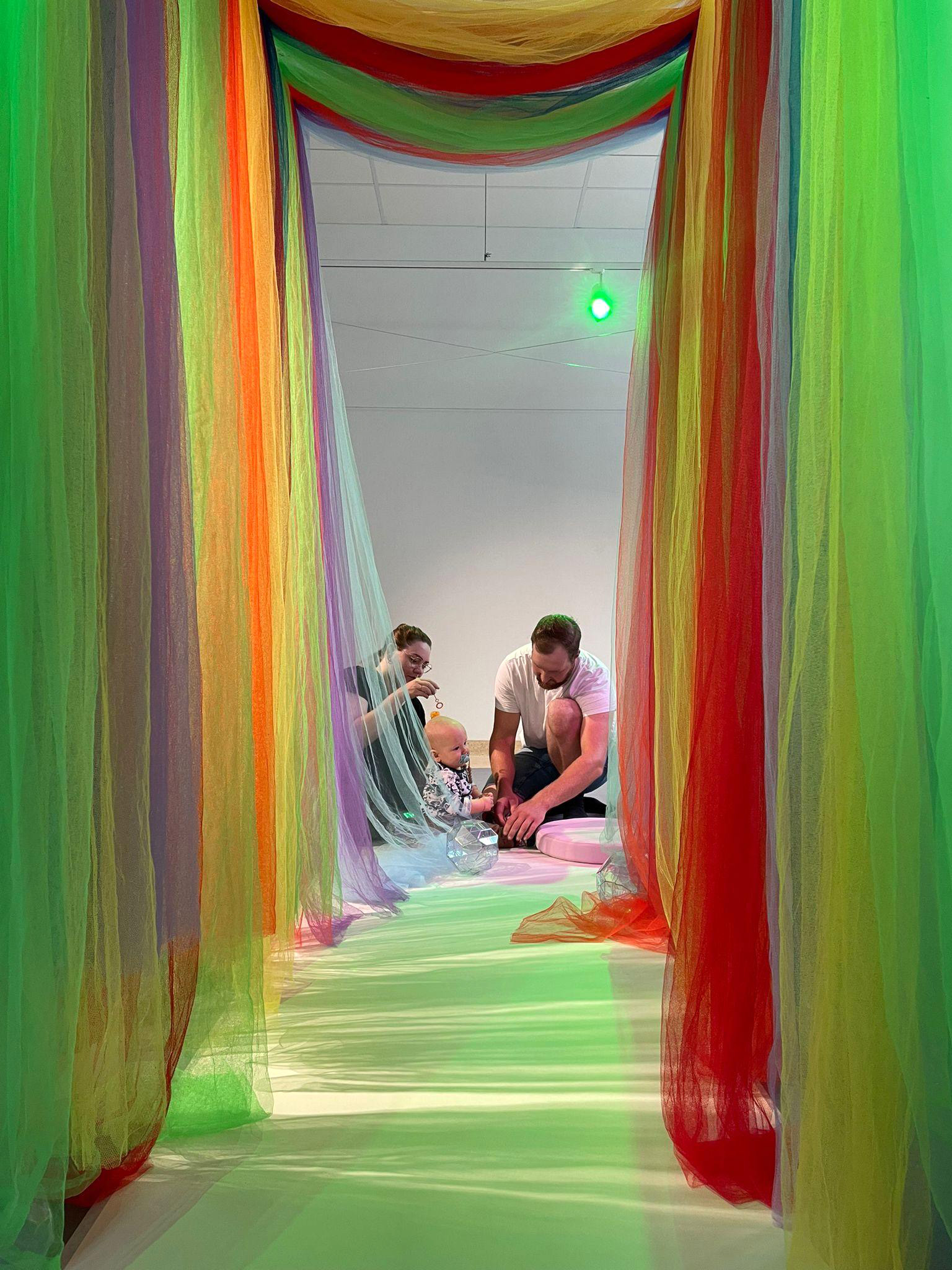 A baby and their parents and carers at the end of a hall filled with colourful materials