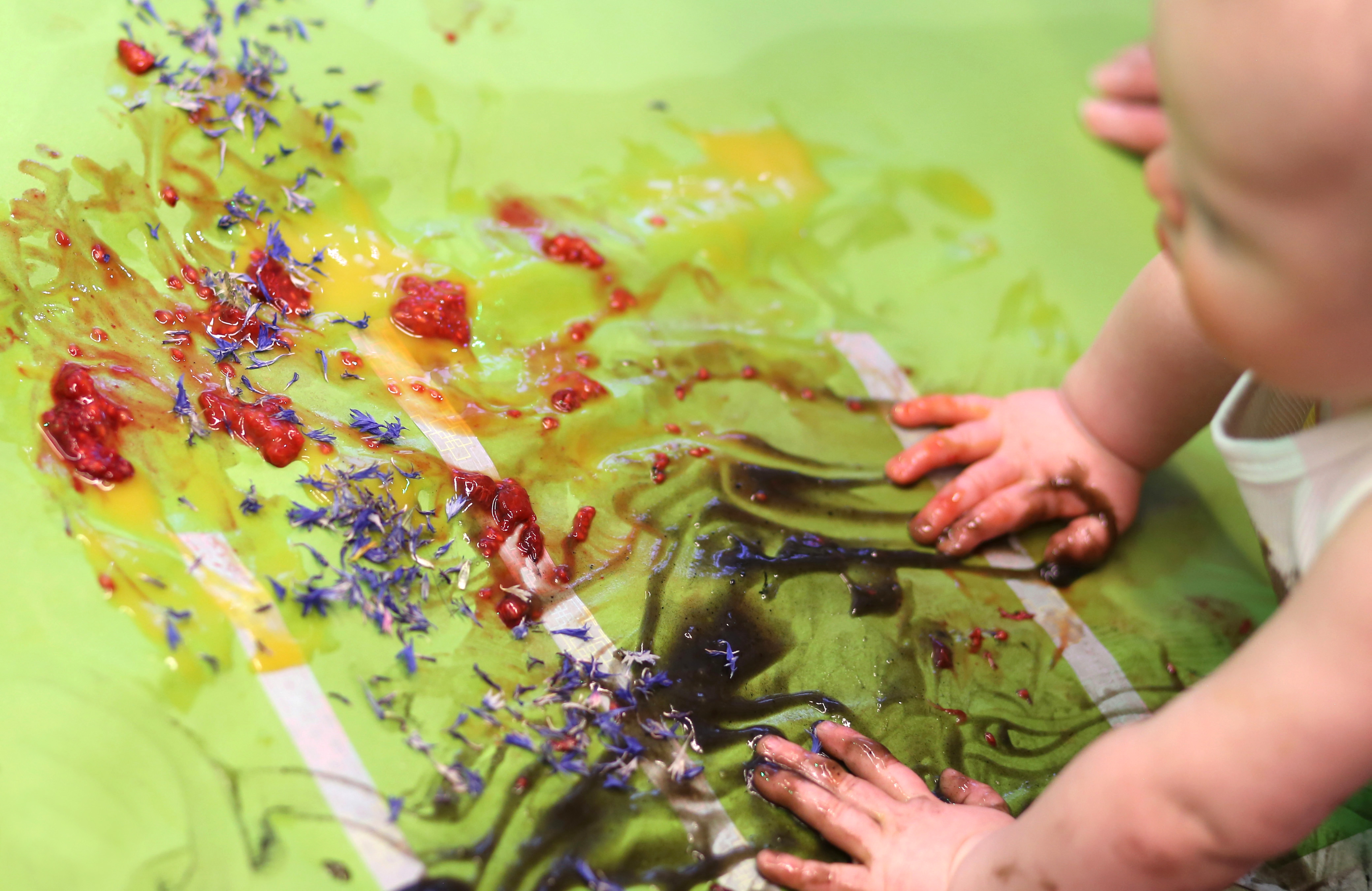 A baby with fruit, flowers and materials on a mat. They have smothered the materials across a green mat. 