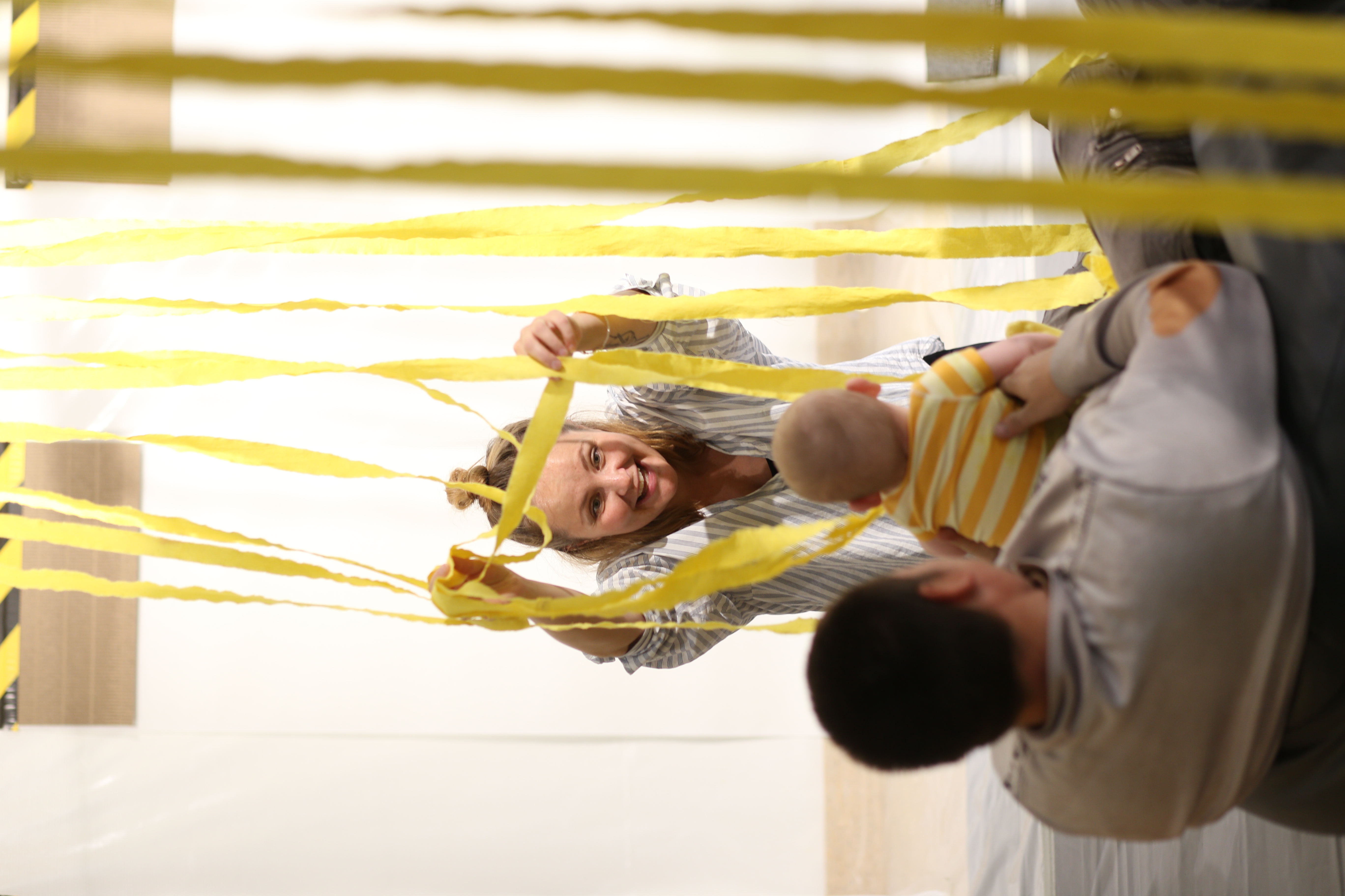 A baby and their parent engaging with an artist holding up yellow string that is hanging from the ceiling 