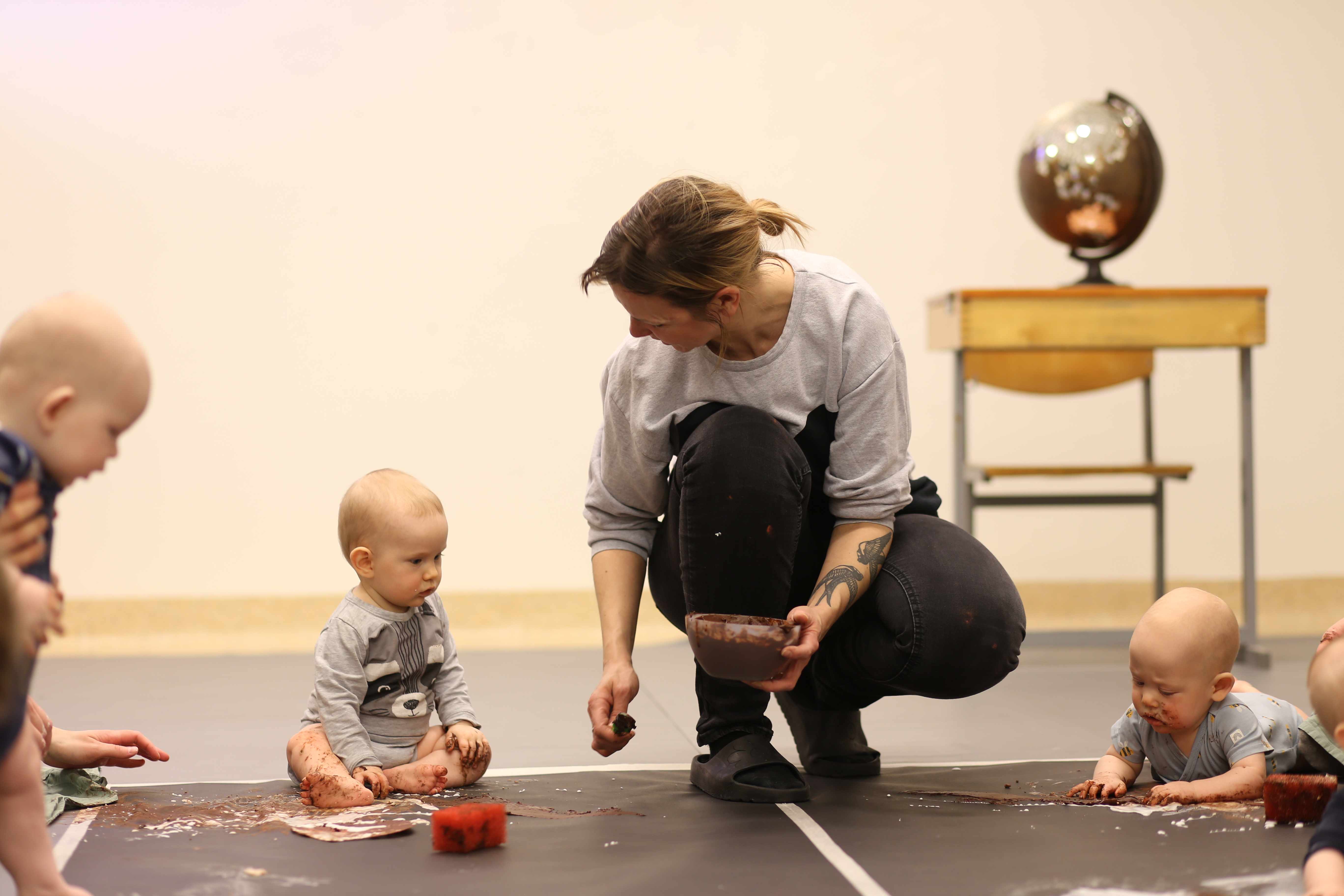A group of babies with an artist who is holding up a box of something. The babies are looking at the artist and on the floor 