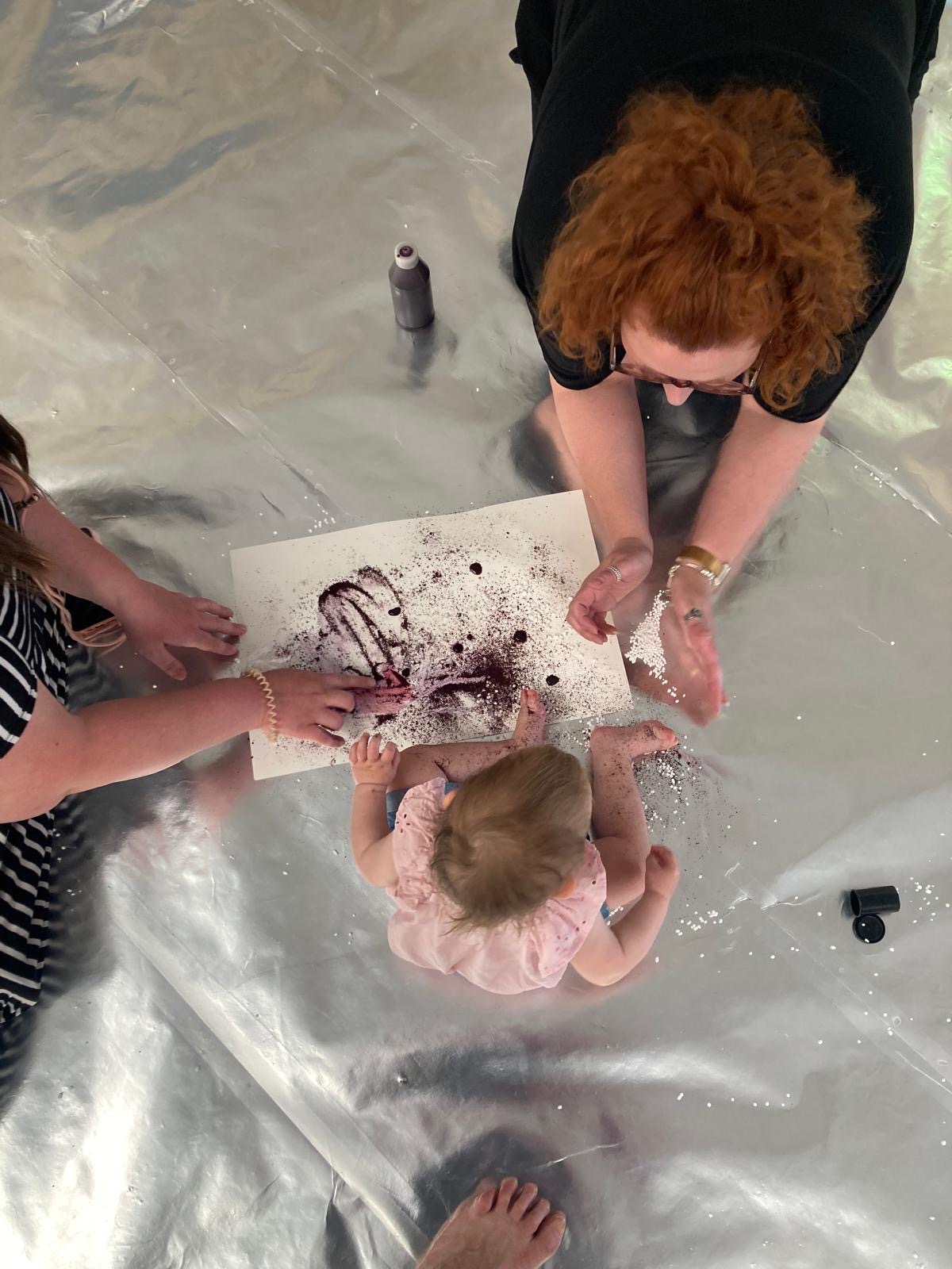 Two adult engage with a young child as they paint using blueberry juice and lemon.