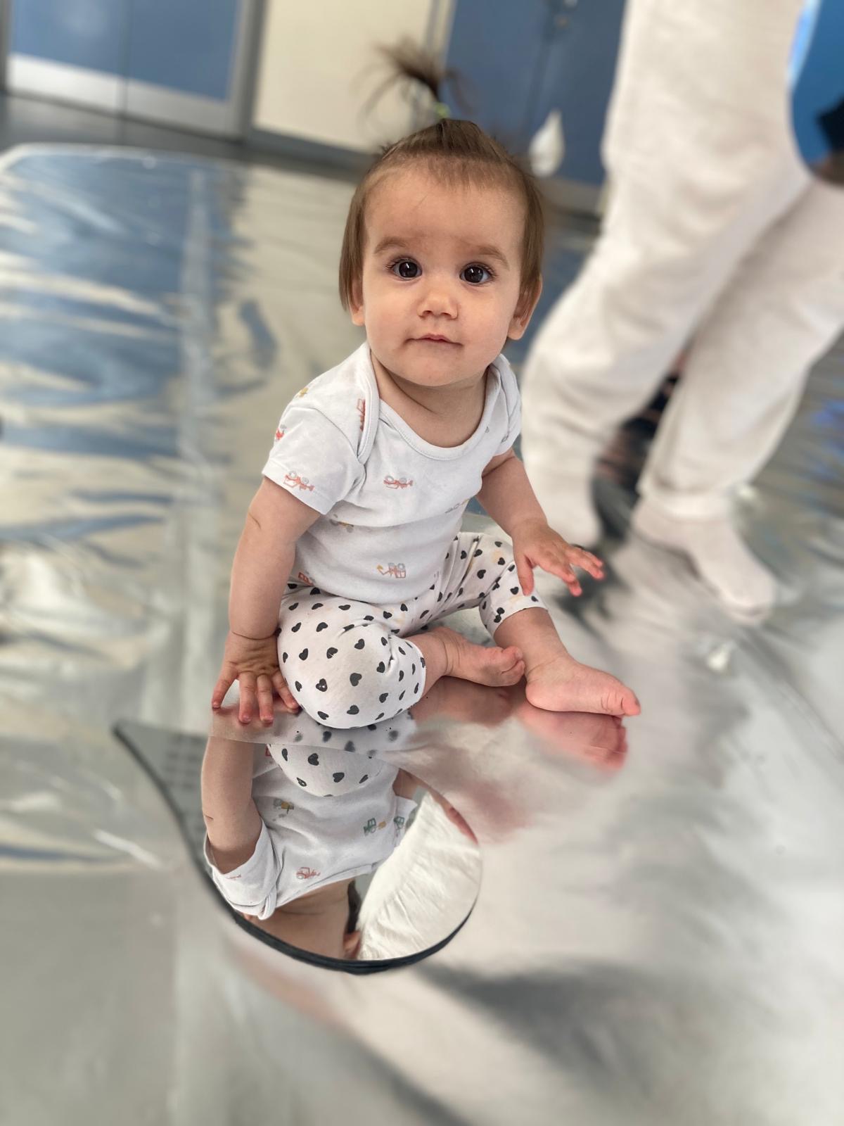 A young child looks at their reflection in a teardrop mirror. The child and the mirror sit on a reflective, silver flooring.