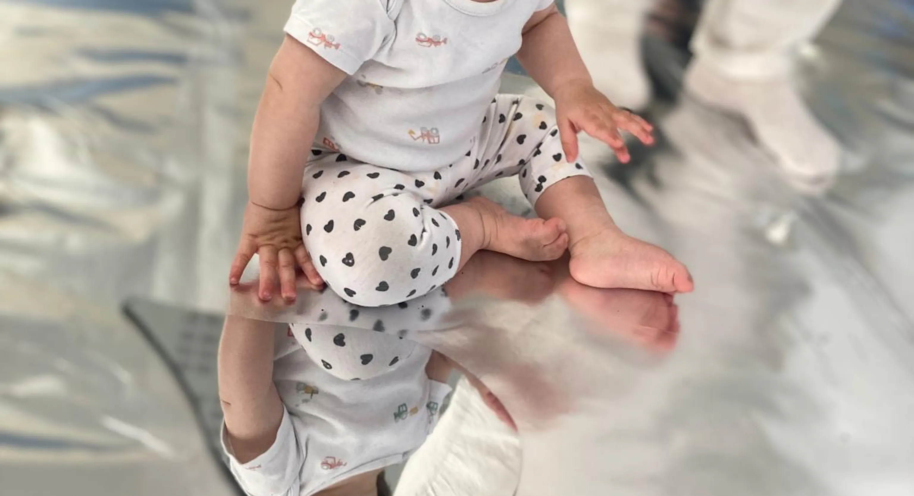 A young child looks at their reflection in a teardrop mirror. The child and the mirror sit on a reflective, silver flooring.