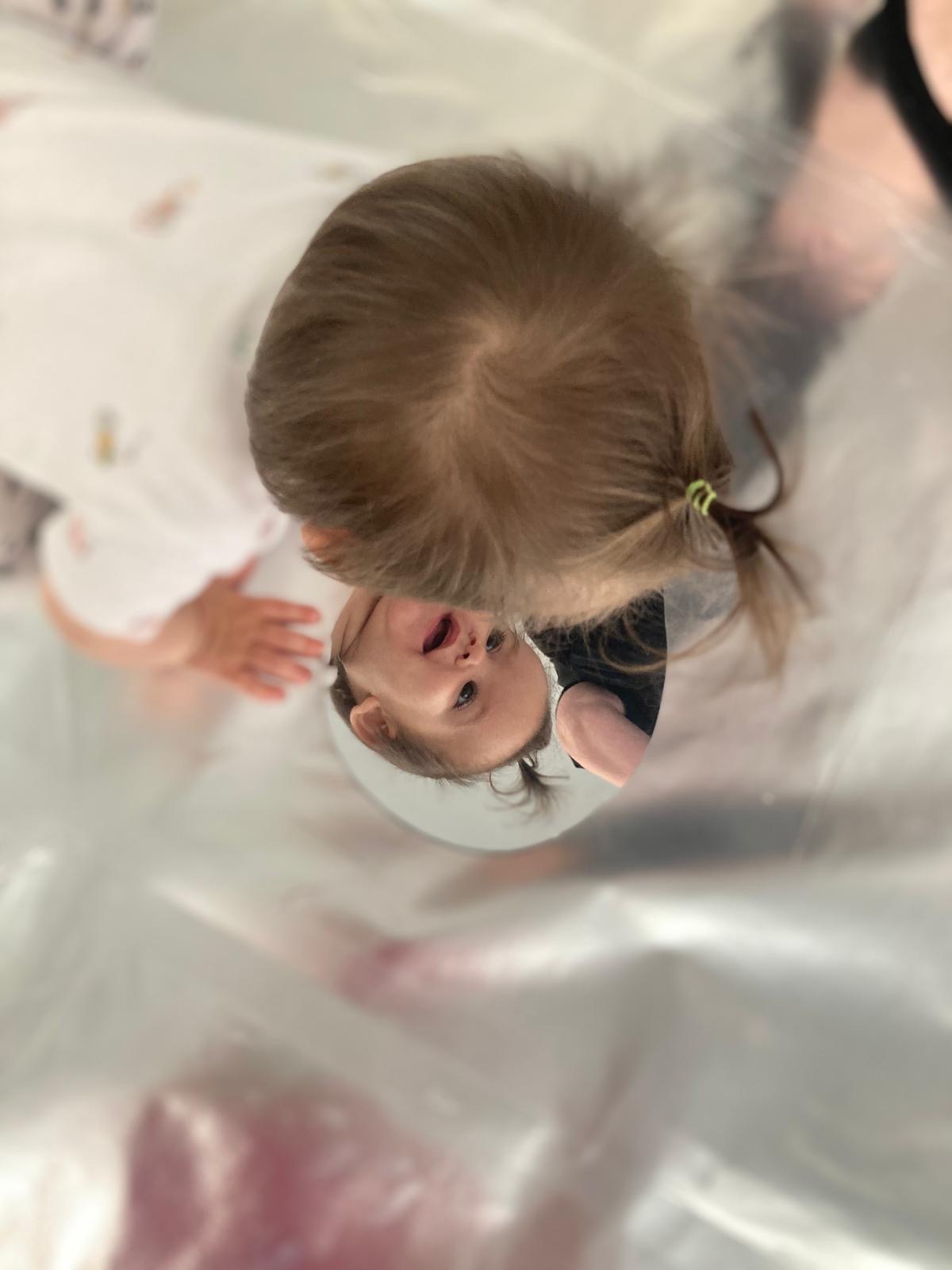 A young child engages with their reflection in a mirror. The mirror and child are on top of silver flooring which reflects the light in the room.