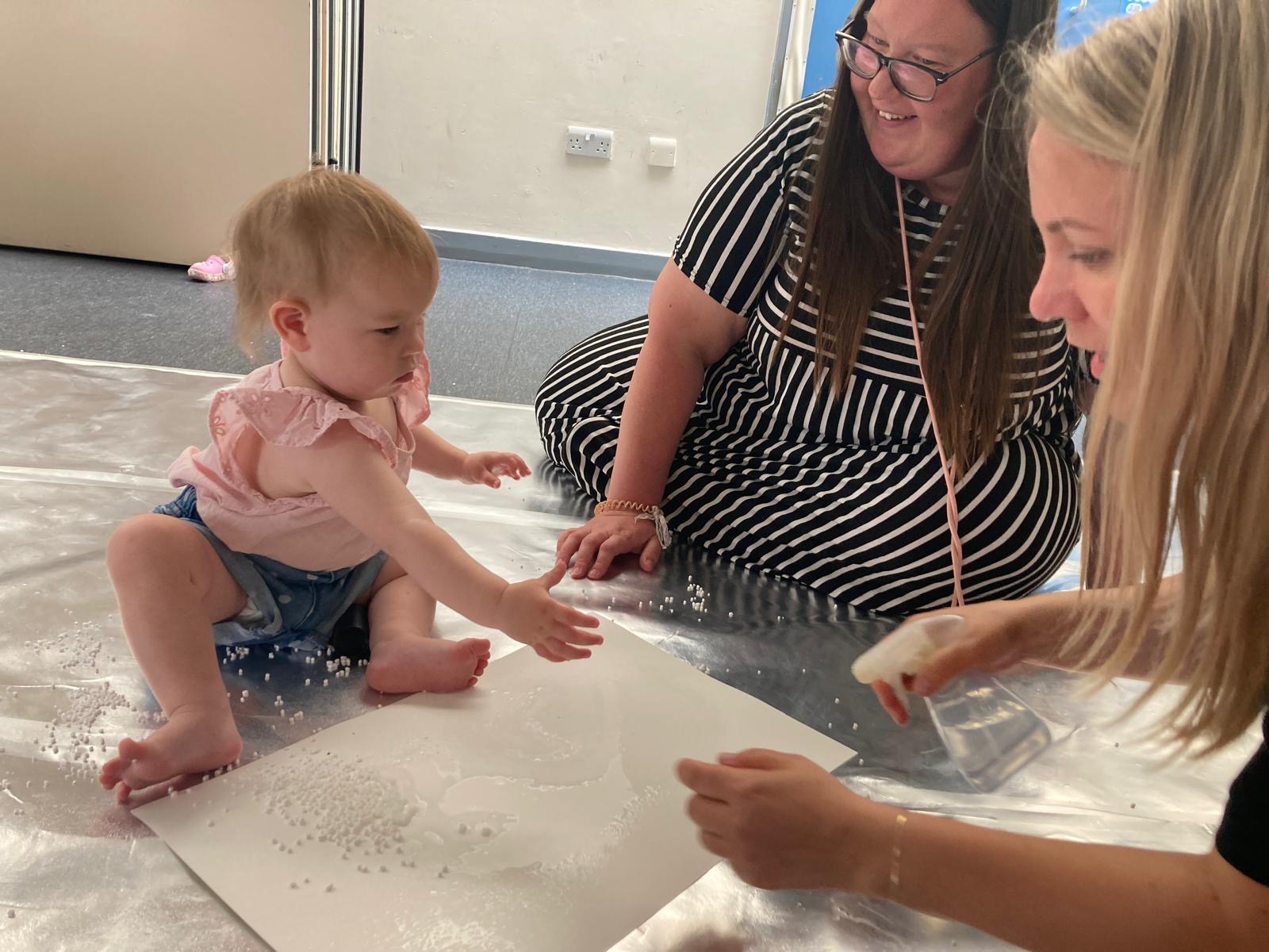 Two adult engage with a young child as they experiment with materials.
