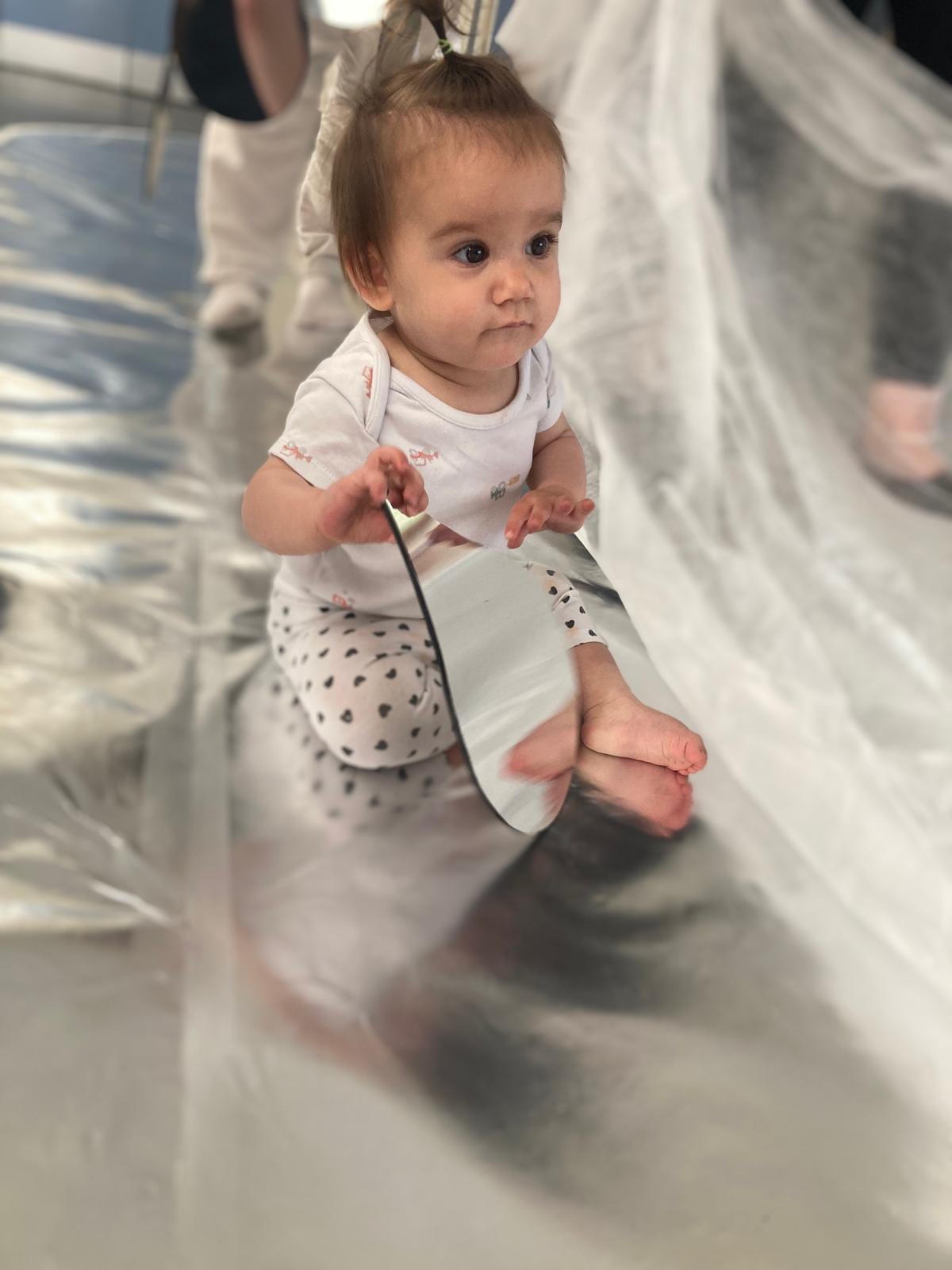 A young child looks at their reflection in a teardrop mirror. The child and the mirror sit on a reflective, silver flooring.