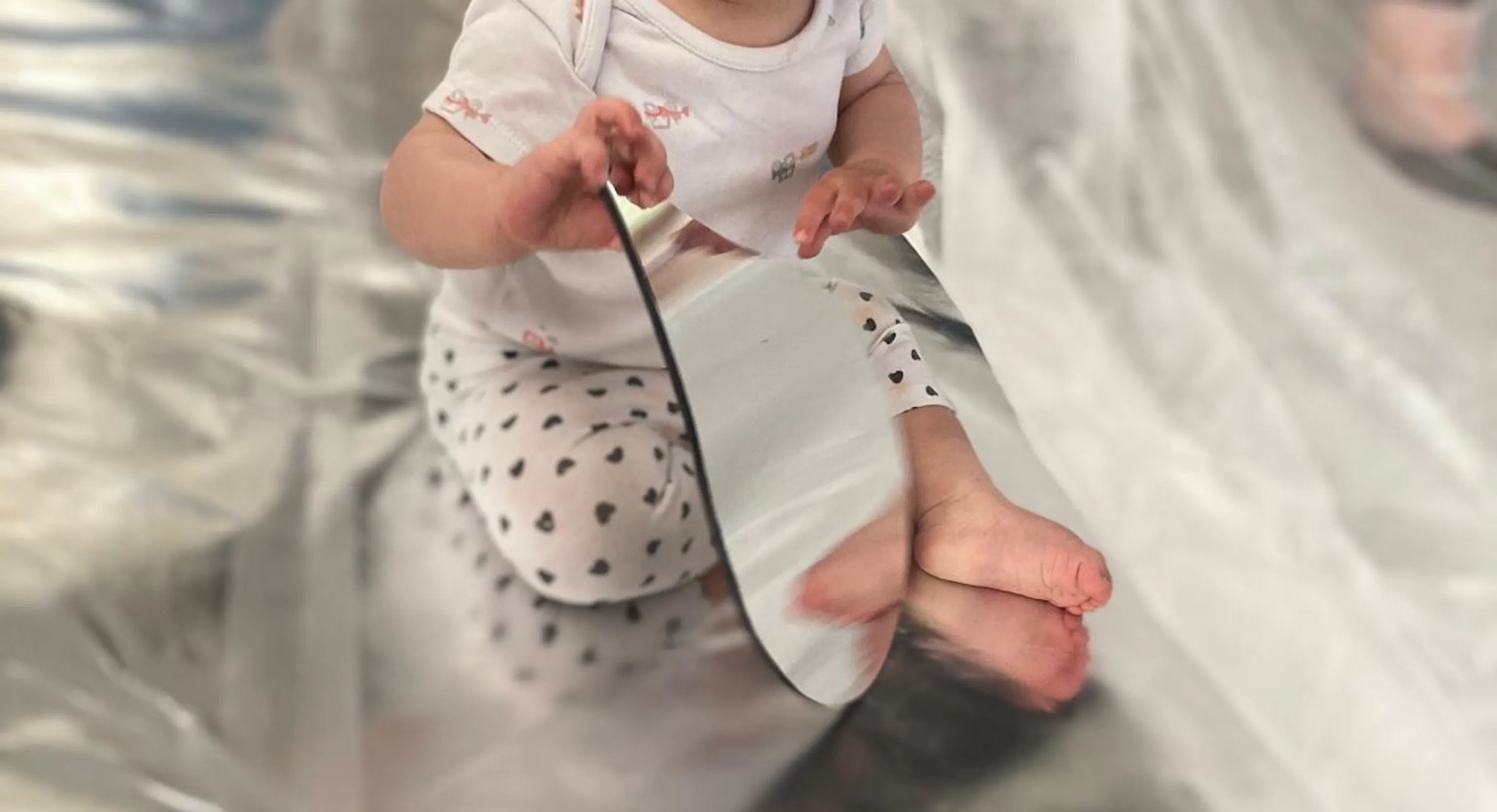 A young child looks at their reflection in a teardrop mirror. The child and the mirror sit on a reflective, silver flooring.