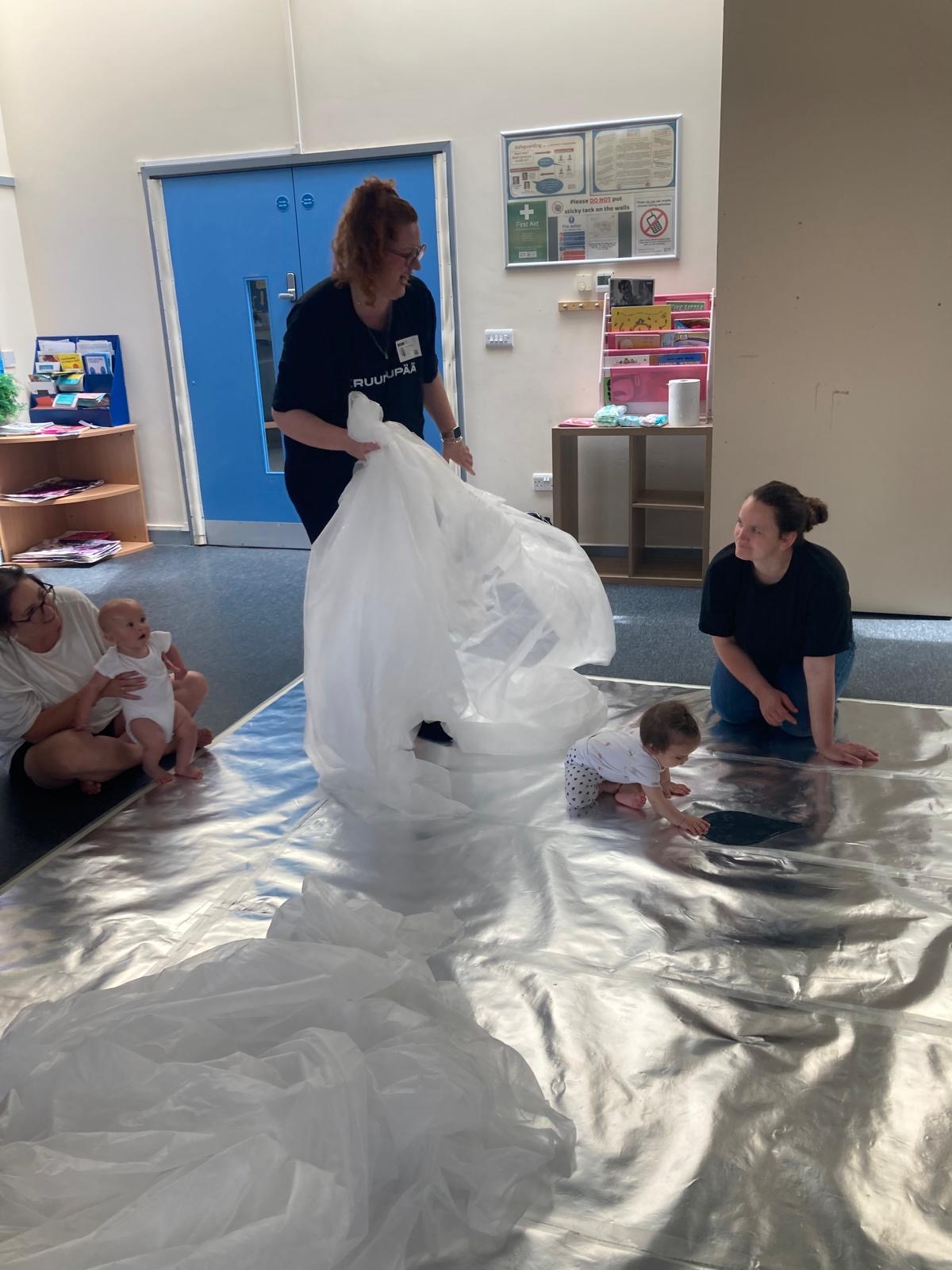 A young child engages with their reflection on a shiny silver floor. There is a large translucent sheet that breaks up the space as the child explores.