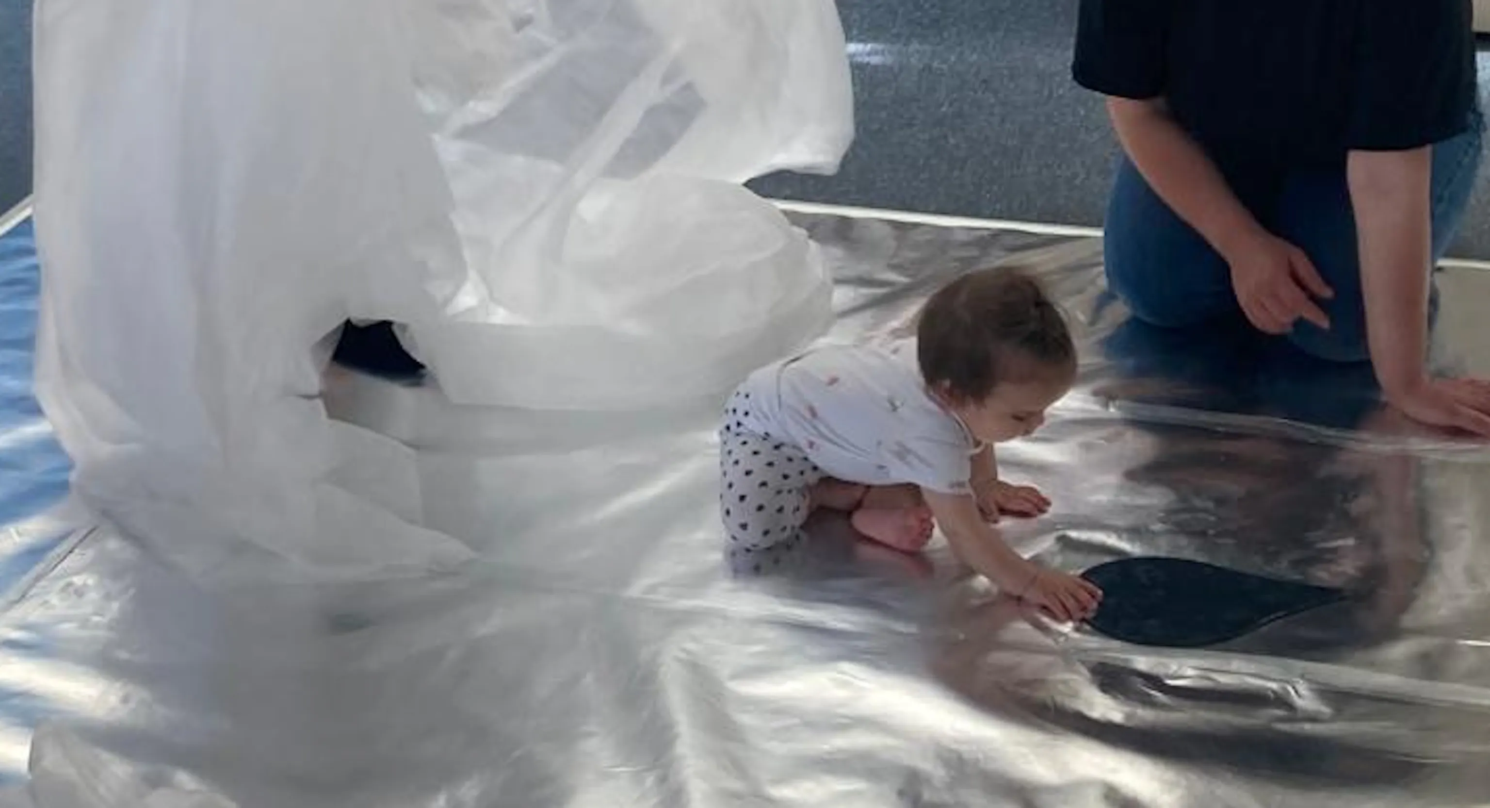 A young child engages with their reflection on a shiny silver floor. There is a large translucent sheet that breaks up the space as the child explores.