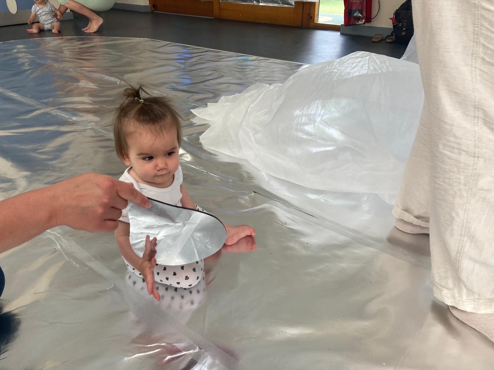 A young child looks at their reflection in a teardrop mirror. They sit on a shiny, silver floor that reflects the light in the room.