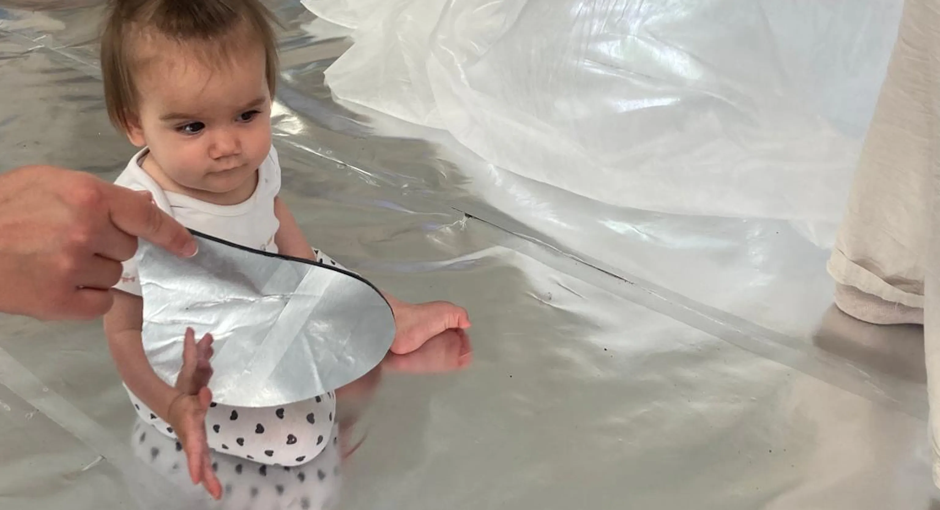 A young child looks at their reflection in a teardrop mirror. They sit on a shiny, silver floor that reflects the light in the room.