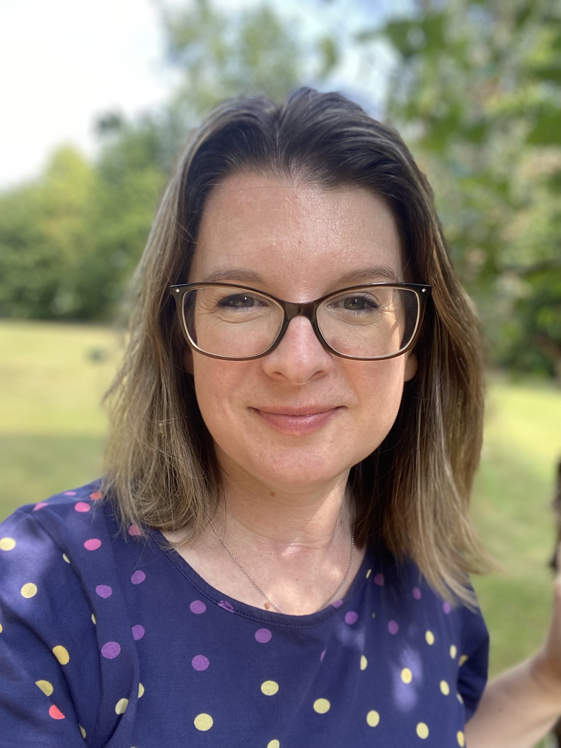 A woman with short blonde/brown hair looking at the camera. In the background are trees and grass. 