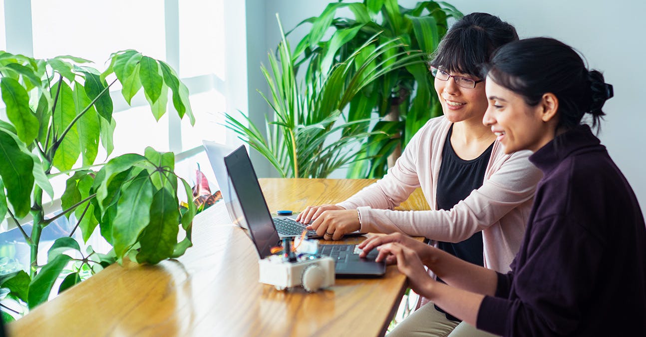 two women at plotly typing on laptops while sitting at desk smiling