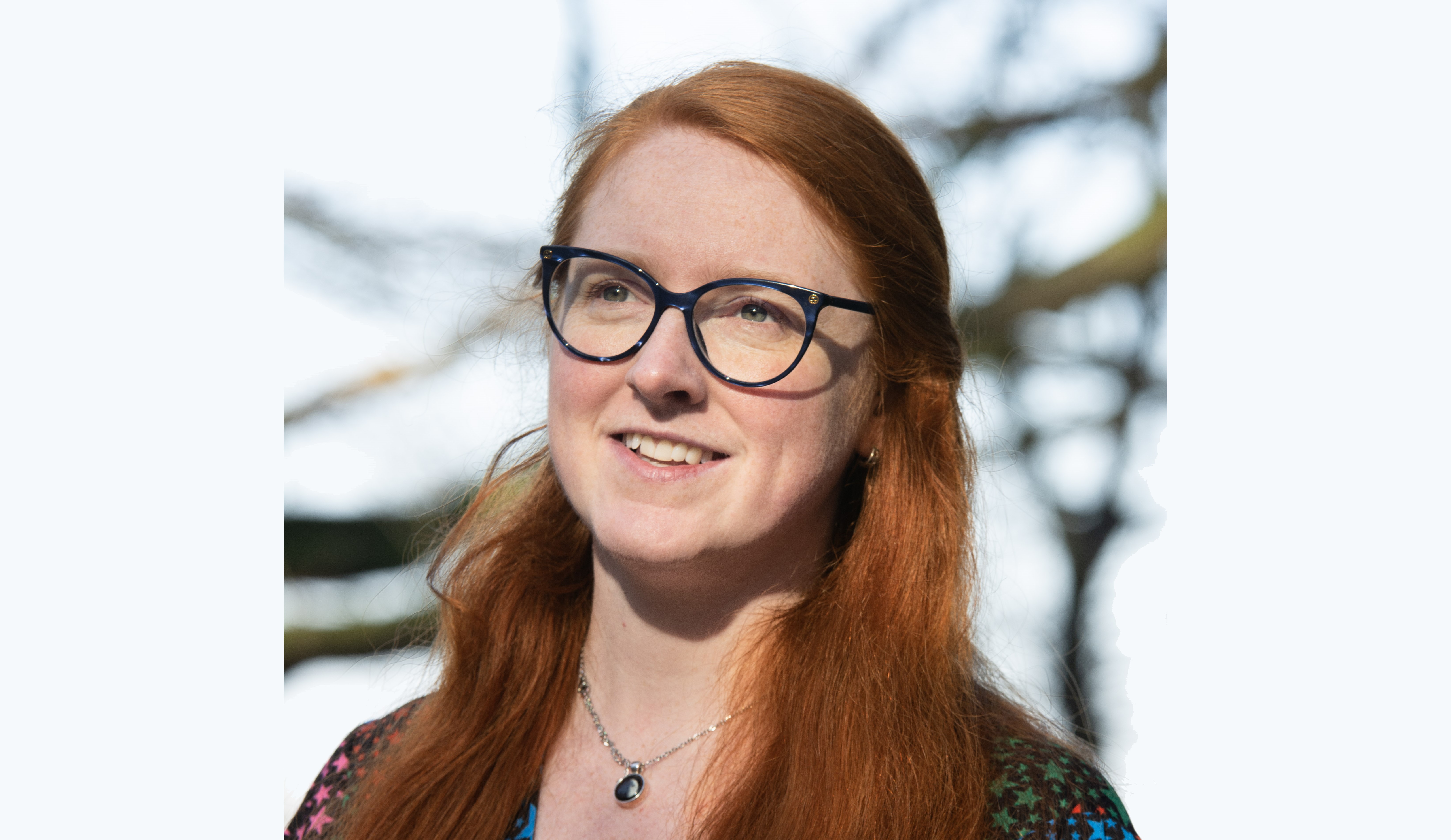 Close up. A woman with red hair and glasses smiling while looking into the distance.