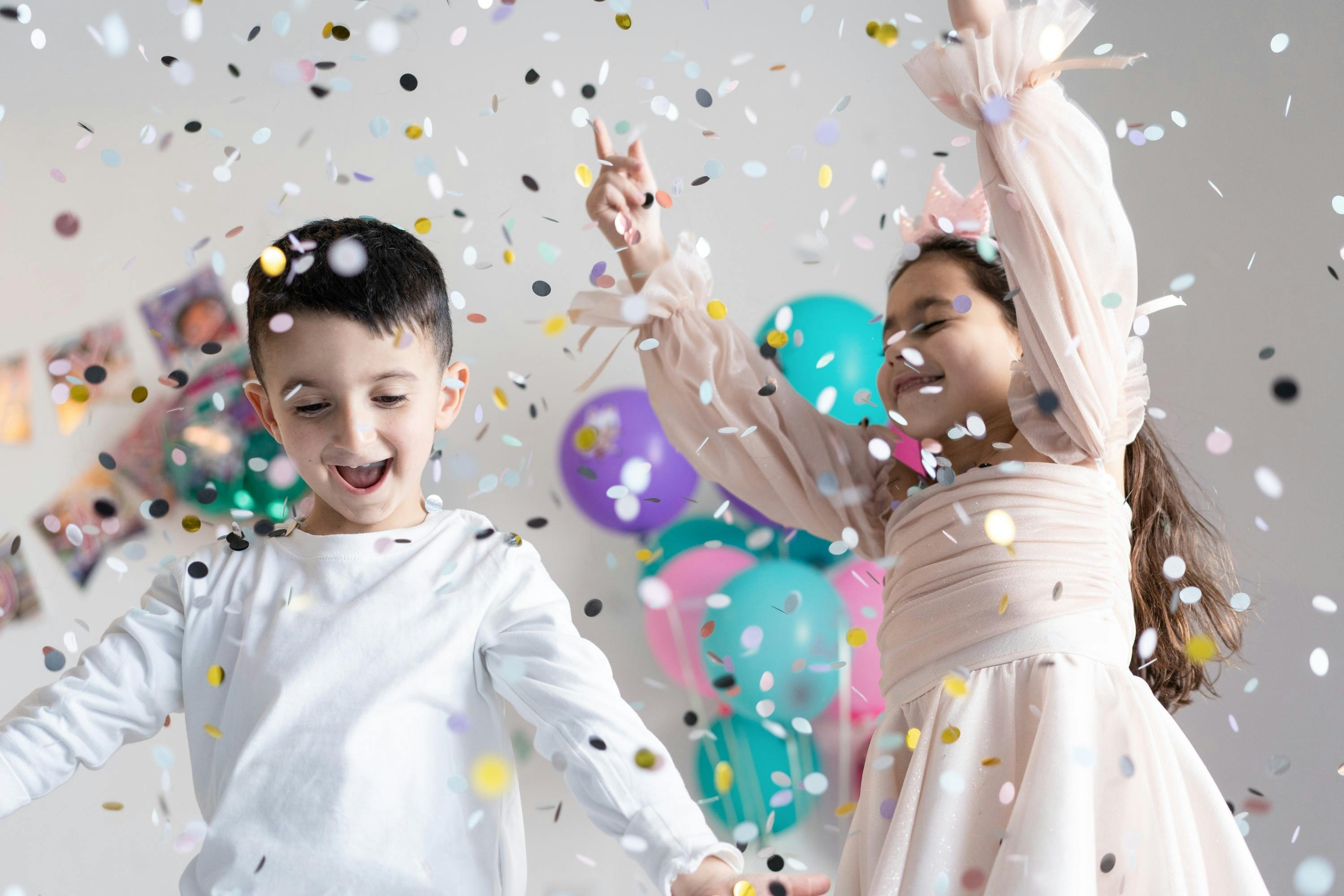 A young boy and girl around 7 years old, dancing happily in white and gold confetti. There are balloons and bunting in the background. The girl wears a pale pink party dress and has her hands in the air with delight. 