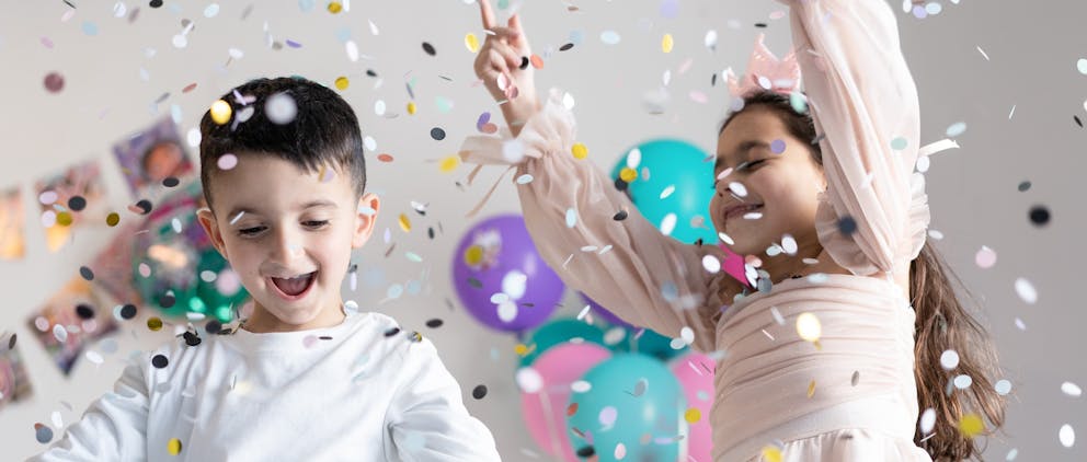 Poppy Walker Children's Celebrant. A boy and girl age 8 are dancing in confetti as it falls down onto them. There are blue and pink balloons and bunting in the background. 