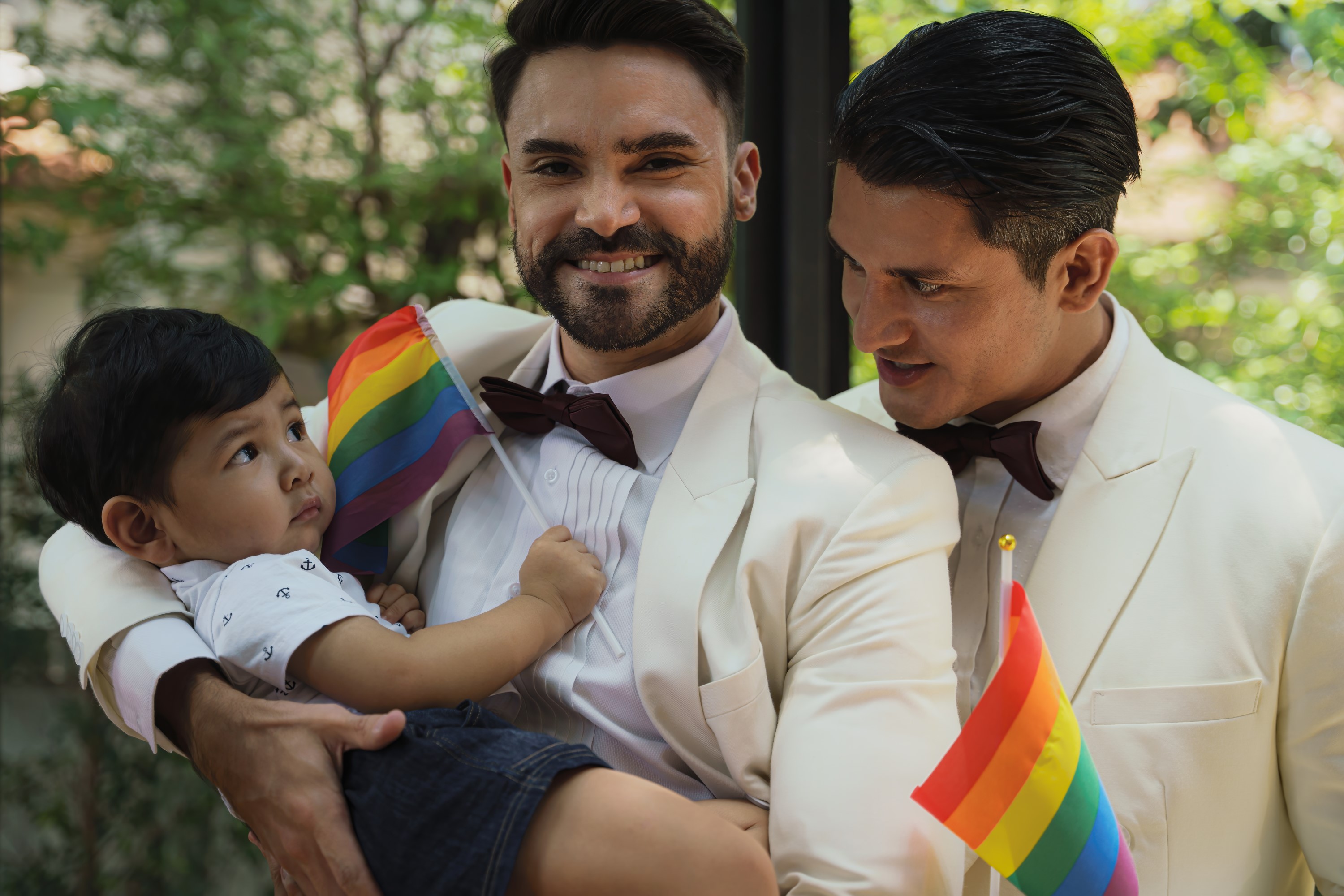 Poppy Walker Children's Celebrant. A gay couple, both wearing white tuxedo's. Cuddle a little boy age 2 who is holding a rainbow flag. The man on the right looks lovingly at the child, the man on the left smiles at the camera.b