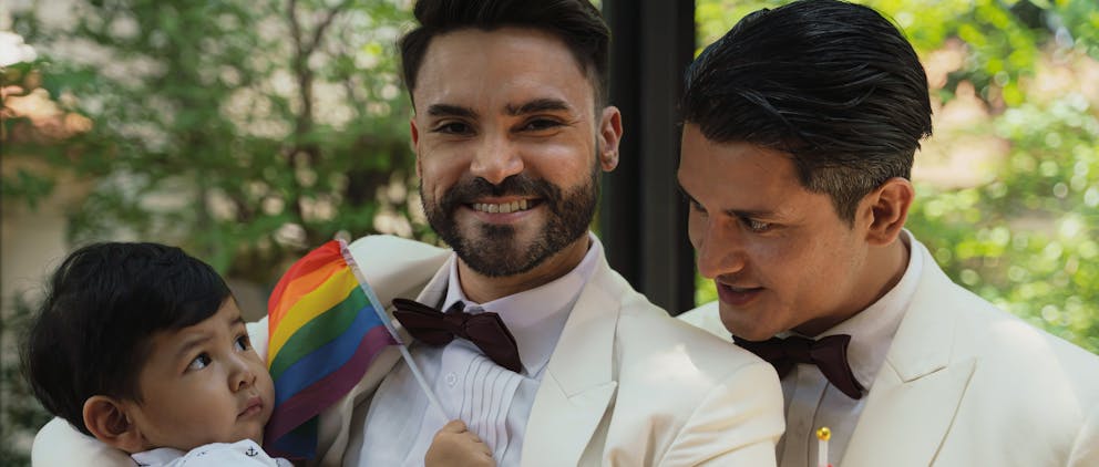 Poppy Walker Children's Celebrant. A gay couple, both wearing white tuxedo's. Cuddle a little boy age 2 who is holding a rainbow flag. The man on the right looks lovingly at the child, the man on the left smiles at the camera.b