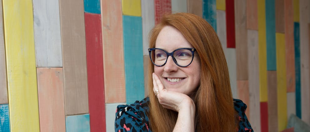 Poppy Walker Children's Celebrant sits against a colourful wooden wall. She has red hair and glasses. Smiling she looks to her right, resting her chin on her hand.