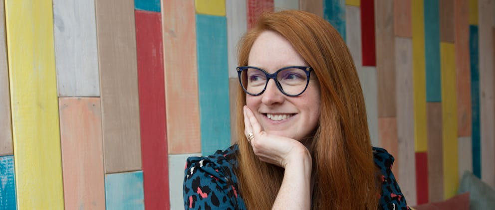 Poppy Walker Children's Celebrant. Against a colourful wooden wall, a headshot of Poppy the celebrant looking over her shoulder, resting her head on her chin. 