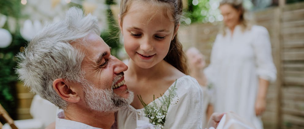 Poppy Walker Children's Celebrant. A man with white hair and a beard is hugging a girl of 10 years old who is say on his knee. They are both smiling and wearing white party clothes. In the background there is a woman and smaller child, both dressed in white, talking and smiling.