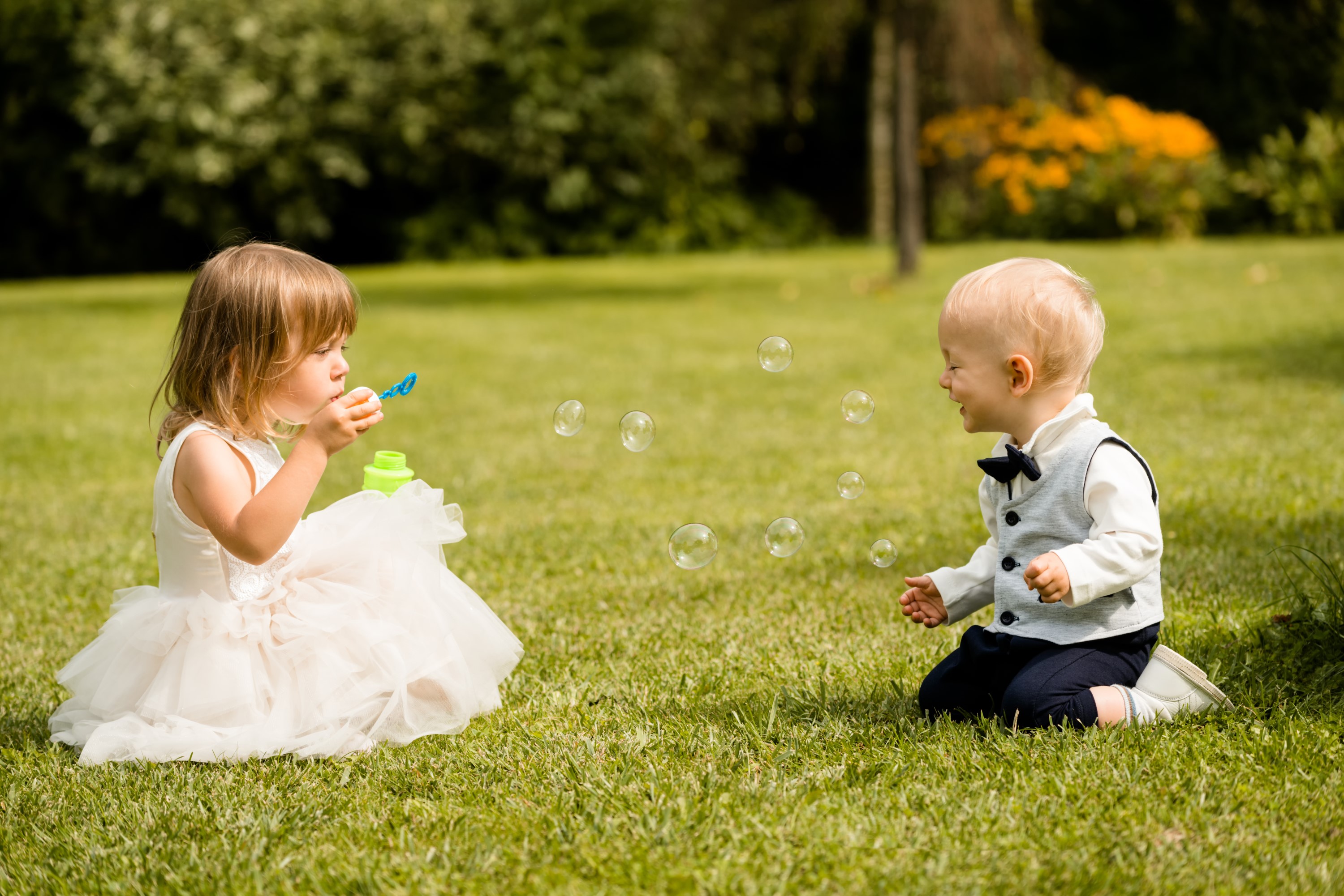 Poppy Walker Children's Celebrant. A little girl in a white party dress sits on green grass. She is blowing bubbles towards a toddler boy in a suit, who is kneeling on the grass and laughing with his eyes closed in delight. 