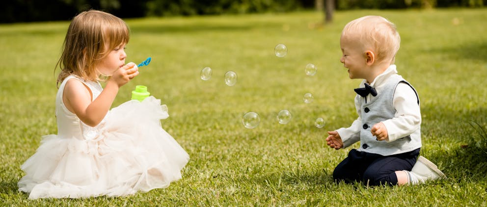 Poppy Walker Children's Celebrant. A little girl in a white party dress sits on green grass. She is blowing bubbles towards a toddler boy in a suit, who is kneeling on the grass and laughing with his eyes closed in delight. 