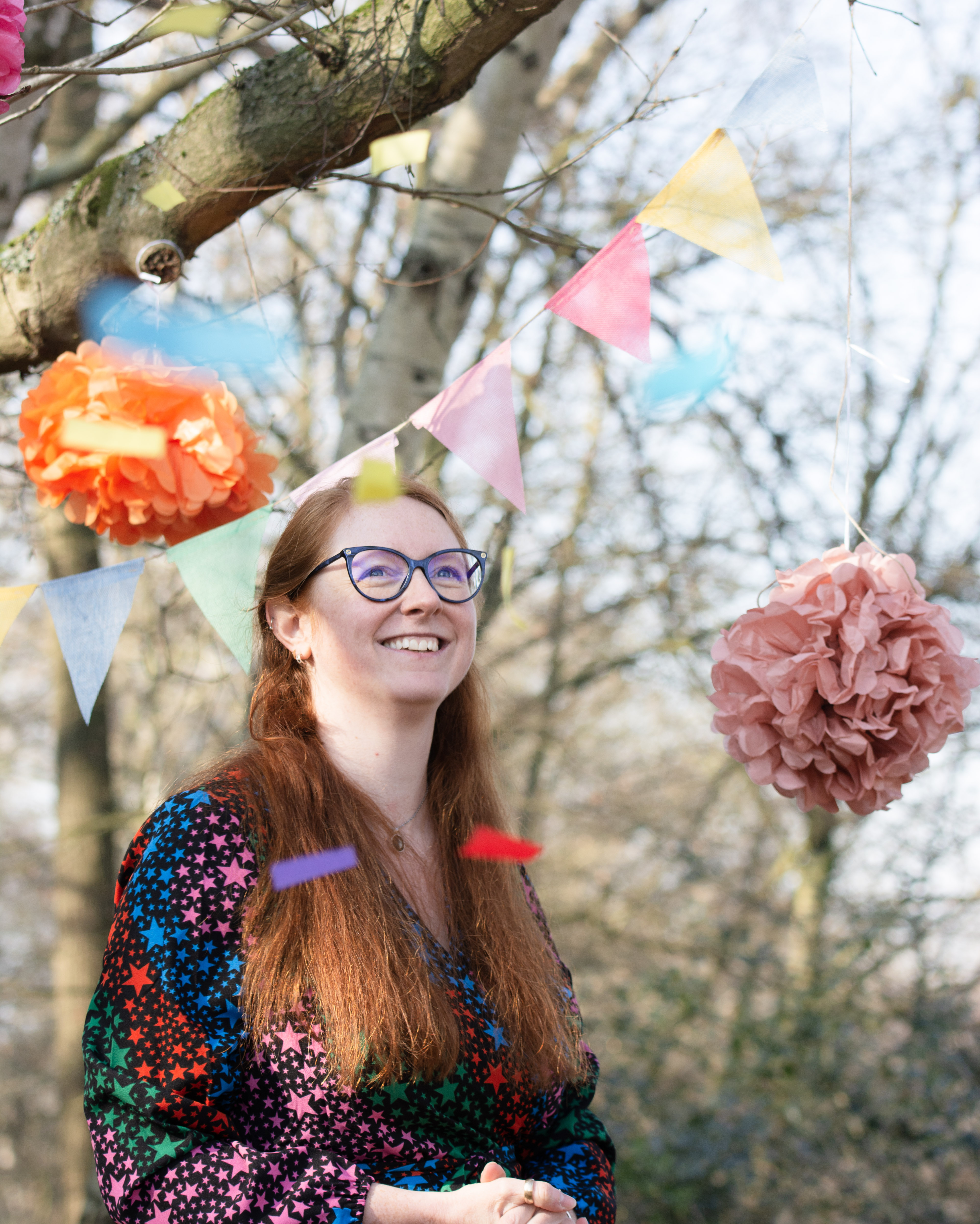 Poppy Walker Children's Celebrant is at an outside at a ceremony, with pink pompoms and pastel bunting in the background. She is smiling as she watches confetti fluttering through the air. 