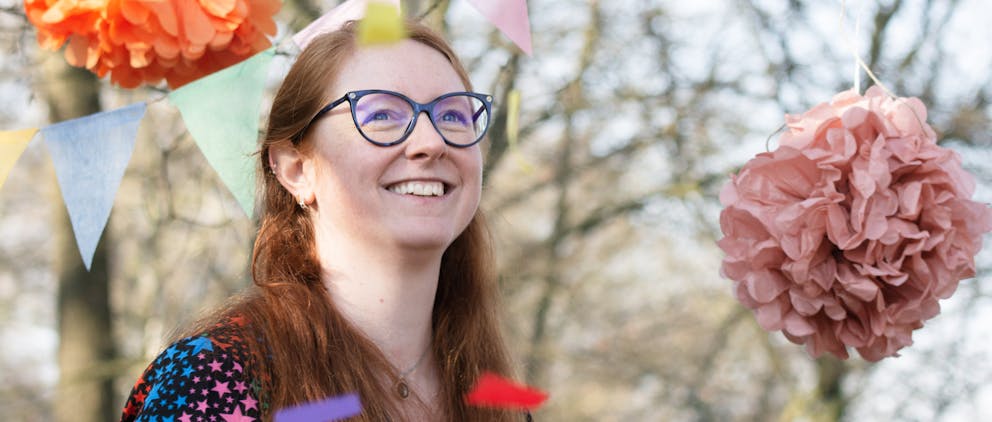 Poppy Walker Children's Celebrant is at an outside at a ceremony, with pink pompoms and pastel bunting in the background. She is smiling as she watches confetti fluttering through the air. 