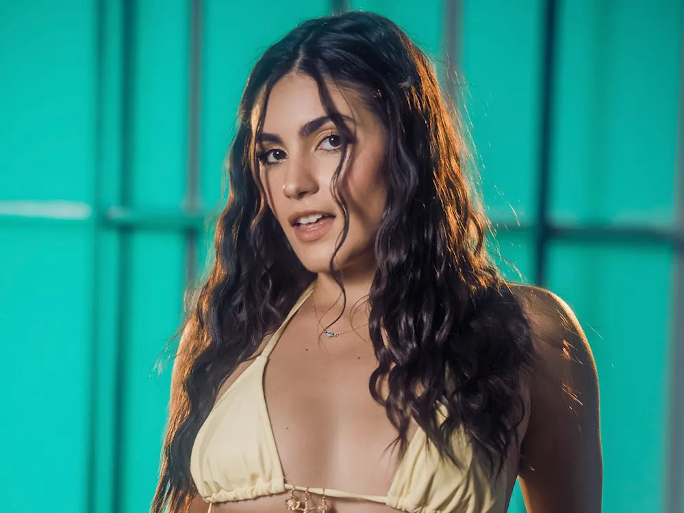 Adult performer Isa Bella poses in a pale-yellow bikini against Dean Capture’s teal backdrop at PornCrush Media Day 2026.