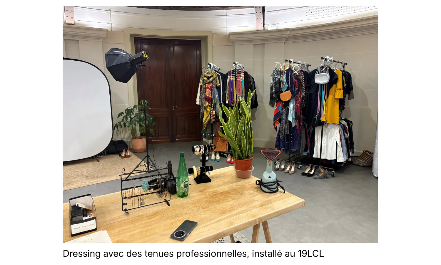 une table en bois en premier plan décoré de plante, porte bijoux, au fond un set up photo et à droite de la photo il y a un dressing