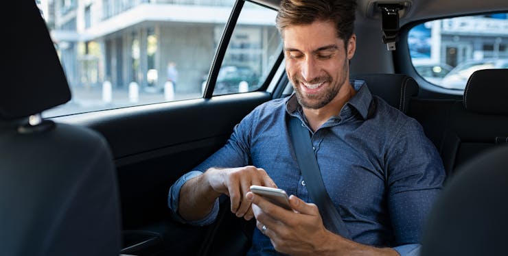 Homem sorrindo e mexendo no celular no banco traseiro de um carro