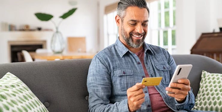 Homem sentado no sofá com celular e cartão de crédito na mão