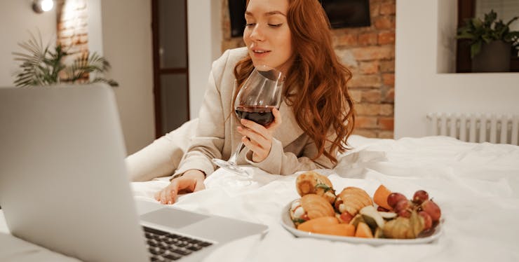 mulher toma vinho enquanto usa o notebook na cama