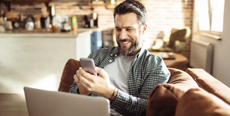 Homem sentado no sofá com um celular e um computador 