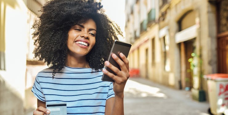 mulher na rua sorrindo com um celular e cartão na mão
