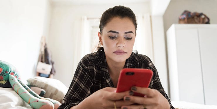 mulher branca de cabelo preto deitada de bruços na cama com o celular na mão