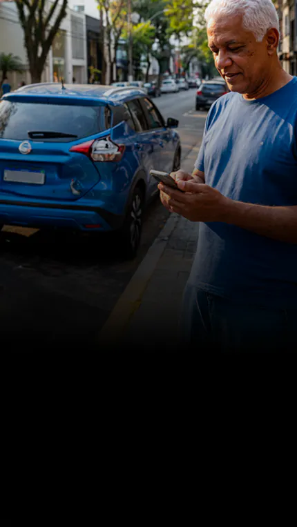 Homem grisalho olha o celular em calçada, ao lado de carro azul com a traseira amassada.