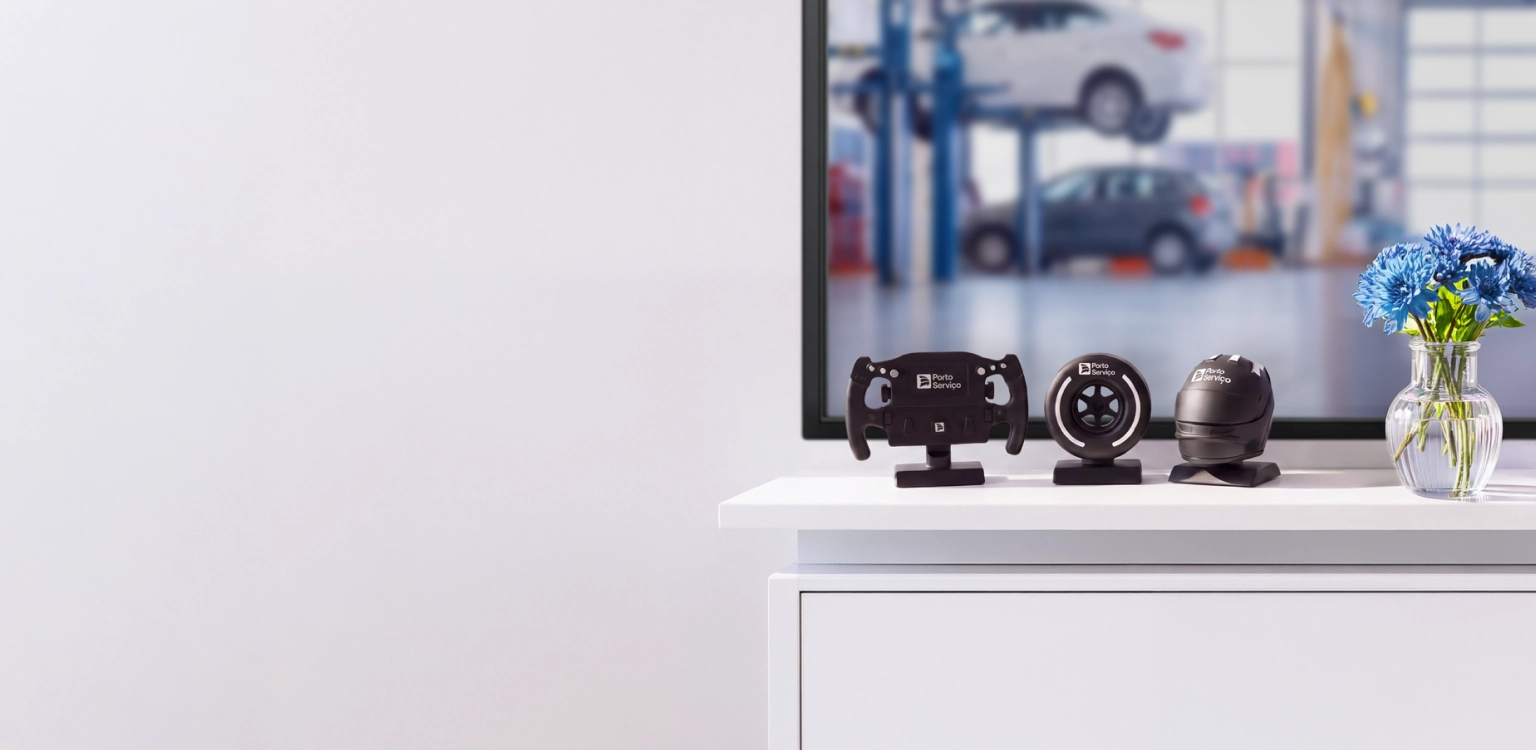Três miniaturas pretas colecionáveis de um capacete de automobilismo um pneu e um volante de carro de corrida com botões, todos com o logotipo branco da Porto Serviço estão expostas sobre um móvel branco, ao lado de um vaso com flores azuis.