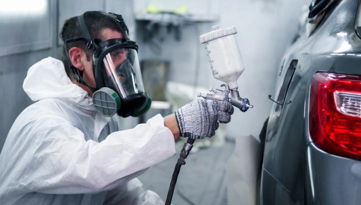 Técnico em traje de proteção completo e máscara respiratória aplicando tinta cinza com uma pistola de pintura na lateral de um carro.