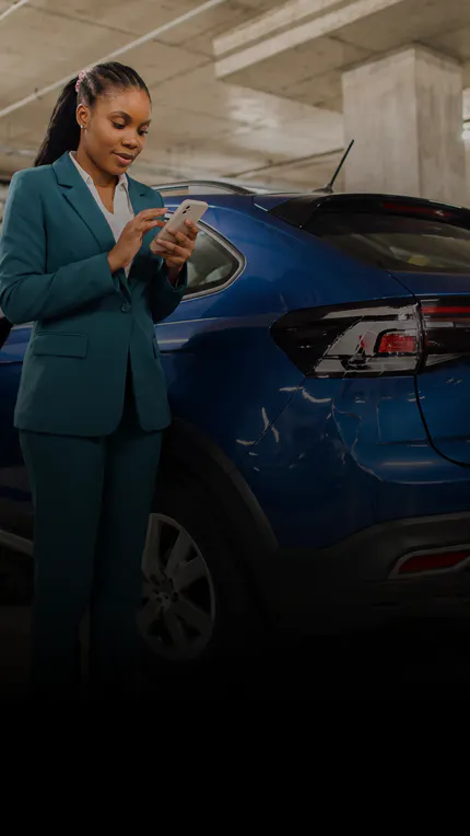 Mulher de terno usa o celular ao lado de carro azul danificado em estacionamento coberto.