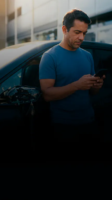 Homem de camiseta azul usa o celular ao lado de um carro com o retrovisor quebrado.