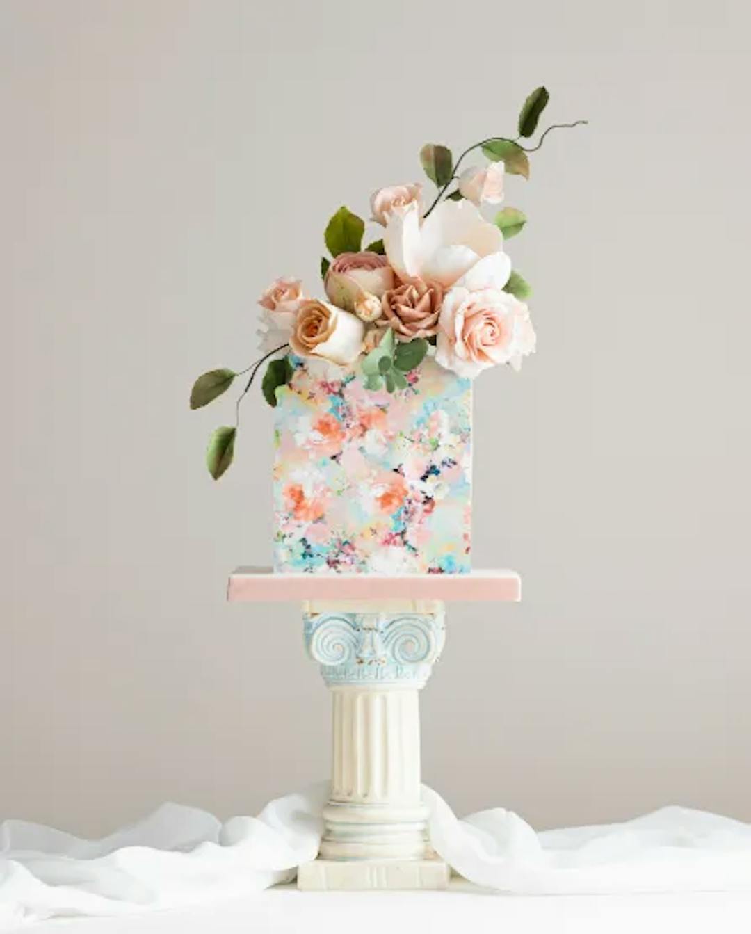 A floral-painted celebration cake with sugar flowers on a Greco-Roman pedestal.