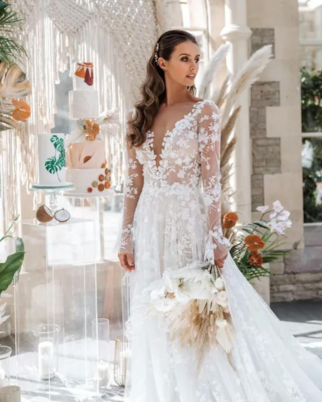 A bride in a lace wedding dress standing beside an elegant boho-style wedding cake display.