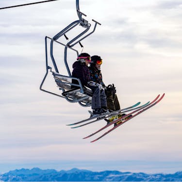 Two skiers ride a chairlift with a sunset and mountains in the background