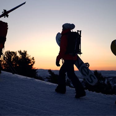One skier and two snowboarders walk uphill with the rising sun while carrying their boards/skis